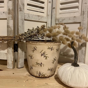 Peut inclure: Un pot en céramique blanc avec un bord brun et un motif de branches d'olivier et de baies. Le pot est rempli de fleurs séchées et est posé sur une surface en bois à côté d'une citrouille blanche.