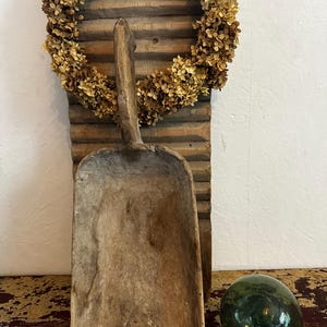 Könnte beinhalten: Eine verwitterte Holzschaufel mit langem Stiel vor einem vertikal ausgerichteten, gerippten Waschbrett aus Holz. Ein kreisförmiger Kranz aus getrockneten goldbraunen Hortensienblüten umgibt den oberen Teil des Waschbretts, und rechts daneben liegt eine dunkelgrüne Glaskugel.