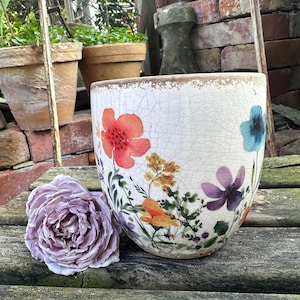Op de afbeelding: Een witte keramieken plantenpot met een craquelé glazuurafwerking en een kleurrijke bloemenprint van rode, oranje, gele, paarse en blauwe wilde bloemen. Een enkele, grote, lichtpaarse kunstbloem ligt naast de plantenpot op een houten ondergrond.
