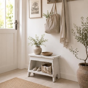 Puede incluir: Un banco de madera blanca con un estante inferior que contiene una cesta tejida y zapatos claros. Encima hay un jarrón de cerámica con vegetación y un cuenco de madera. Un bolso de tela beige y una tela cuelgan de un perchero de pared de madera.