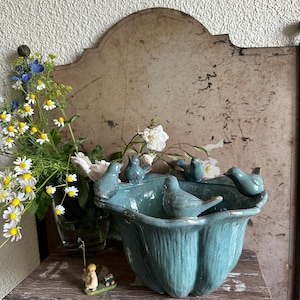 Puede incluir: Un bebedero de cerámica turquesa con forma de tulipán, adornado con varias figuritas de pájaros pequeños alrededor del borde. El bebedero se exhibe sobre una superficie de madera, con una pequeña figura de una persona y flores en el fondo.