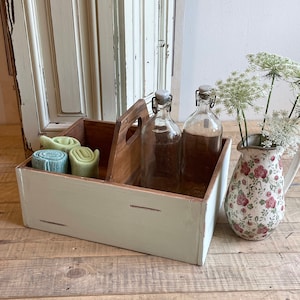 Può includere: Una scatola di legno con esterno verde chiaro e interno in legno naturale. Due bottiglie di vetro trasparente con chiusure a scatto all'interno. Panni verdi e blu arrotolati in un vano. Una brocca floreale con fiori bianchi a destra.