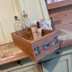 Op de afbeelding: Een houten lade met een metalen handvat met het label "35". De lade is gevuld met glazen flessen en een bundel grijze kaarsen. De lade staat op een lichtgroen houten oppervlak.