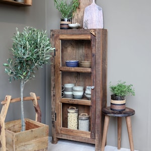 Puede incluir: Un armario rústico de madera con puertas de cristal exhibe varios artículos de cocina. En el interior hay tazas, cuencos y tarros llenos de pasta y cereales. Una pequeña planta en maceta se encuentra sobre un taburete de madera.