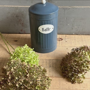 Könnte beinhalten: Eine blaue Metall-Kaffeedose mit Deckel und weißem Knopf. Die Dose hat ein weißes ovales Etikett mit der Aufschrift "Kaffe". Die Dose steht auf einer Holzoberfläche mit grünen und braunen Trockenblumen.