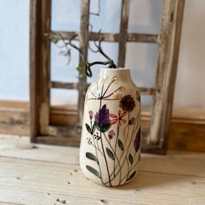 Può includere: Un vaso in ceramica bianca con un design floreale con fiori viola, rosa, arancioni e gialli. Il vaso ha un collo stretto e un corpo arrotondato.