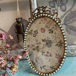 Peut inclure: Cadre photo ovale en or avec une bordure de perles et un insert à motif floral. Le cadre est orné d'un nœud décoratif en haut.