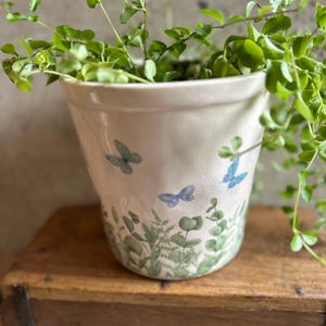 Pode incluir: Um vaso de cerâmica branca com um acabamento de esmalte craquelado, decorado com um padrão de folhas de eucalipto verdes e pequenas borboletas azuis à volta da base e dos lados, contendo uma planta verde pendente de folhas pequenas.