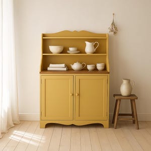 Könnte beinhalten: Ein senfgelbes Sideboard mit offenen Regalen und einem zweitürigen Unterteil. Die Regale zeigen weißes Geschirr, darunter Schalen, Teller, eine Teekanne und eine Kanne. Ein kleiner Holzschemel hält eine passende Kanne.