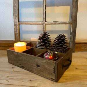 May include: A rustic wooden box with compartments, holding a lit candle, pine cones, and a red ornament. The box has a dark finish and a metal handle. A weathered window frame is in the background, creating a vintage aesthetic.