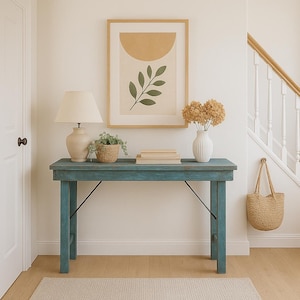 May include: A rectangular blue console table with a distressed finish and black metal support braces. On the table sits a beige lamp, a small potted plant, two stacked books, and a white vase filled with dried flowers. A woven bag hangs nearby.