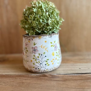 Pode incluir: Um pequeno vaso de cerâmica branco com um padrão floral, segurando um arranjo de flores verdes secas. O vaso tem uma aparência envelhecida e desgastada com uma borda marrom. O design floral inclui flores rosa, folhas verdes e detalhes amarelos. O vaso está em uma superfície de madeira.