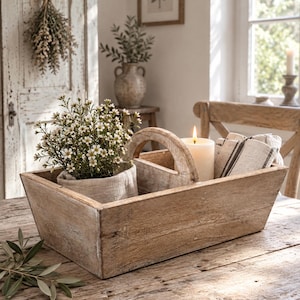 Op de afbeelding: Een rechthoekige houten mand met een centrale boogvormige handgreep en twee vakken. De ene kant bevat een plant met kleine witte bloemen in een beige stoffen wikkel, de andere kant een brandende witte stompkaars en gevouwen gestreepte stof.