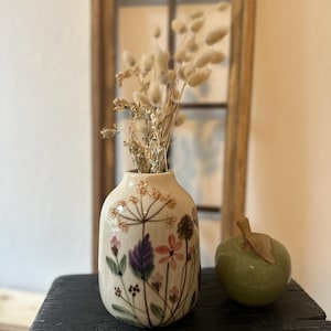 Könnte beinhalten: Eine cremefarbene Keramikvase mit floralen Designs in Rosa, Lila und Braun, gefüllt mit getrockneten beigen Blumen. Ein grüner Keramikapfel mit braunem Stiel steht neben der Vase auf einer schwarzen Oberfläche.