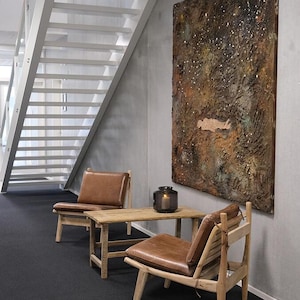 May include: Two brown leather chairs with wooden frames and a wooden table with a brown leather cushion. A dark grey lantern sits on the table. A large abstract painting with brown, gold, and white colours hangs on the wall.