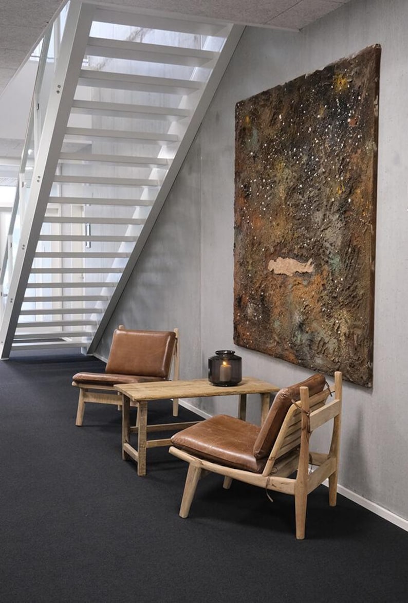 May include: Two brown leather chairs with wooden frames and a wooden table with a brown leather cushion. A dark grey lantern sits on the table. A large abstract painting with brown, gold, and white colours hangs on the wall.