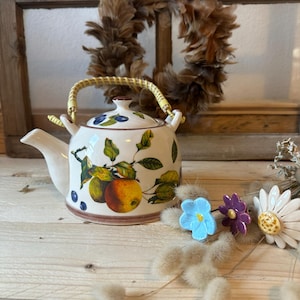 Può includere: Una teiera in ceramica bianca con un manico giallo e un design floreale con mele, foglie e mirtilli. La teiera è appoggiata su una superficie di legno con un bouquet di fiori in ceramica.