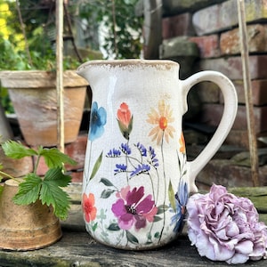 Könnte beinhalten: Ein cremefarbener Keramikkrug mit einer rissigen Glasuroberfläche, verziert mit einem bunten Blumenmuster aus verschiedenen Wildblumen in Blau, Orange, Violett und Pink mit grünen Stielen und Blättern sowie einem geschwungenen Henkel an der Seite.