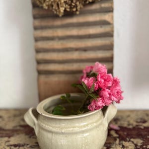 Może przedstawiać: Kremowy ceramiczny garnek z dwoma bocznymi uchwytami, w którym znajduje się bukiet żywych różowych róż. Za garnkiem stoi wysoka, prostokątna drewniana tarka do prania, zwieńczona kompozycją z suszonych kwiatów w warstwowej kompozycji.