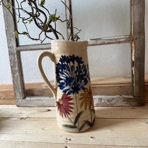 May include: A ceramic pitcher with a floral design. The pitcher is cream colored with blue, red, and yellow flowers. The pitcher has a handle and a spout.