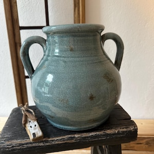 Op de afbeelding: Een turquoise keramische vaas met twee handvatten en een craquelé glazuur. De vaas staat op een kleine, verweerde houten kruk. Naast de vaas staat een klein, decoratief houten huisje.