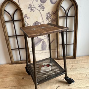 Op de afbeelding: Een rustieke, rollende bijzettafel met een verweerd houten blad en een onderste metalen plank. De tafel heeft een donker metalen frame en zwarte zwenkwielen. Decoratieve boogvormige raamkozijnen en botanische prints zijn op de achtergrond.