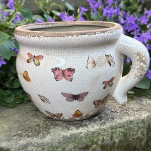 Op de afbeelding: Een witte keramische plantenpot met een verweerde afwerking en een handvat. De pot is versierd met een vlindermotief in tinten roze, bruin en geel.