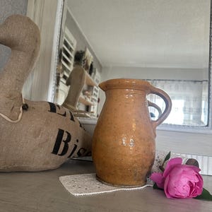 Könnte beinhalten: Eine rustikale, hellbraune Keramikkrug mit Henkel, auf einer weißen Spitzendecke. Eine rosa Rose und eine Gänsefigur aus Jute sind in der Nähe. Der Krug hat eine gesprenkelte Textur und einen leicht ausgestellten Rand.