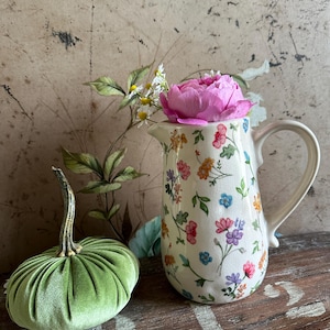 Könnte beinhalten: Ein dekoratives Stillleben mit einem grünen Samtkürbis und einem floralen Krug. Der Krug ist mit bunten Blumen verziert und enthält eine rosa Pfingstrose. Die Gegenstände sind auf einer rustikalen Holzoberfläche angeordnet.
