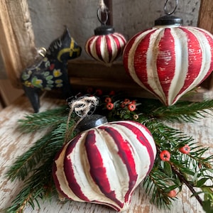 Pode incluir: Dois ornamentos de vidro canelados em forma de cebola com um padrão de riscas vermelhas e brancas desgastadas, tampas de metal preto e cordéis para pendurar. Um ornamento semelhante mais pequeno pende acima, junto a uma figura de cavalo preta.
