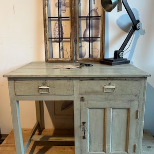 Könnte beinhalten: Ein hellgrüner Holzschreibtisch mit zwei Schubladen und einer Schranktür. Der Schreibtisch hat ein schlichtes Design mit einer flachen Oberseite und geraden Beinen. Der Schreibtisch steht auf einem Holzboden.