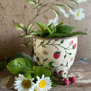 Puede incluir: Una maceta de cerámica con diseño de fresas y flores, llena de menta verde. Cerca hay margaritas y adornos de fresas de ganchillo. La maceta tiene un acabado craquelado y un borde dorado. El fondo presenta un mural floral.