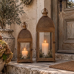 May include: Two rectangular metal lanterns with a weathered bronze finish, featuring arched tops with mesh detailing and glass panels. Each lantern contains a white pillar candle surrounded by green foliage, resting on a stone step.