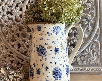 May include: A vintage-style white pitcher with a blue floral pattern, holding dried green hydrangeas. The pitcher has a distressed finish and a handle. The background features a carved wooden panel. A rustic, farmhouse-style decorative piece.