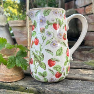 Könnte beinhalten: Ein Keramikkrug mit cremefarbener Basis und einer rissigen Glasuroberfläche. Die Oberfläche ist mit einem botanischen Muster aus roten Erdbeeren, grünen Blättern und kleinen hellvioletten Blüten verziert. Er hat einen geschwungenen Henkel und Ausguss.