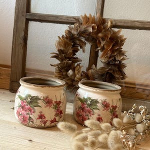 Könnte beinhalten: Zwei cremefarbene Keramiktöpfe mit einem Blumendesign mit rosa Blüten und grünen Blättern. Ein brauner Federkranz und getrocknete Pflanzen befinden sich im Hintergrund. Die Töpfe stehen auf einer Holzoberfläche.
