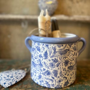 Könnte beinhalten: Eine Keramik-Tasse mit blauem Rand und Henkeln, verziert mit einem blau-weißen Blumen- und Hasenmuster. Die Tasse enthält Kerzen und Nagellack. Ein passendes Etikett liegt daneben.