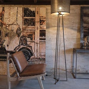 May include: A brown leather chair with a wooden frame sits in a room with a metal floor lamp with a wire mesh shade. The lamp is on and the light is shining on the chair. The wall behind the chair is painted white and has a large picture of a vintage car on it.