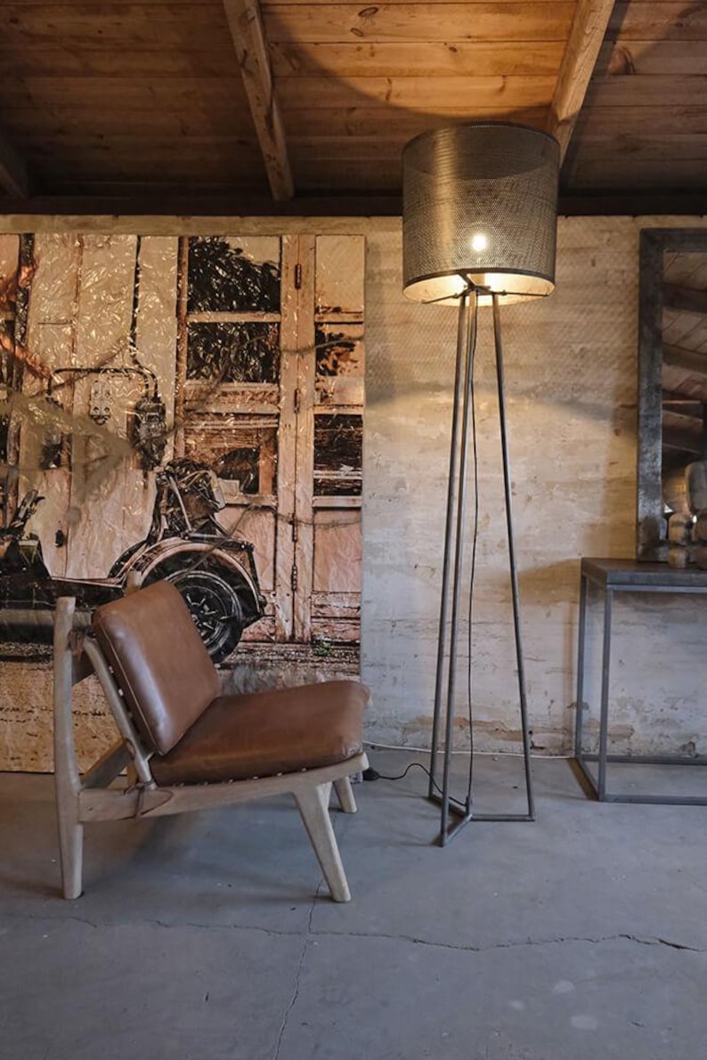 May include: A brown leather chair with a wooden frame sits in a room with a metal floor lamp with a wire mesh shade. The lamp is on and the light is shining on the chair. The wall behind the chair is painted white and has a large picture of a vintage car on it.
