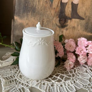 Könnte beinhalten: Weiße Keramikdose mit Deckel, verziert mit einem zarten floralen Muster am Rand. Die Dose steht auf einer weißen Häkelunterlage.