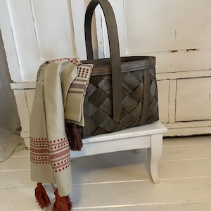 Peut inclure: Un panier en osier brun avec une poignée en bois posé sur un tabouret blanc. Un tissu rayé beige et rouge avec des glands pend sur le côté du panier.