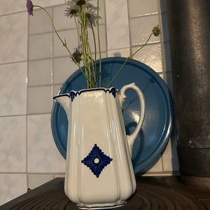 Könnte beinhalten: Eine weiße Keramikkanne mit einem blauen und weißen Diamantenmuster. Die Kanne hat einen Henkel und ist mit weißen und violetten Blumen gefüllt.