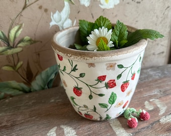 May include: A small, off-white ceramic pot with a strawberry and vine pattern. The pot contains green leaves and a white daisy. A few small, crocheted strawberries sit beside the pot on a wooden surface.