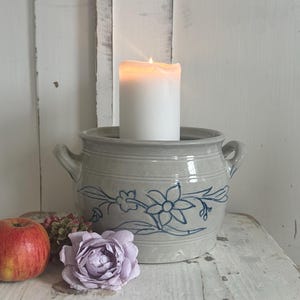 Op de afbeelding: Een brandende witte pilaar kaars staat in een grijze keramische pot met blauwe bloemmotieven en twee handvatten. Een appel en een paarse roos staan naast de pot op een wit houten oppervlak.