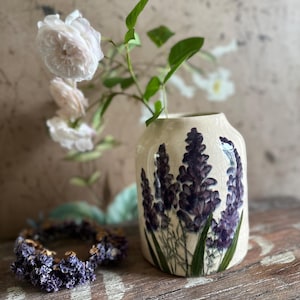 Puede incluir: Un jarrón de cerámica color crema con un diseño floral de lavanda, con rosas blancas y hojas verdes. Una corona de lavanda descansa sobre una superficie de madera. El jarrón tiene un acabado craquelado.