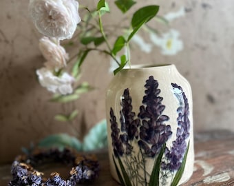 Può includere: Un vaso in ceramica color crema con un motivo floreale di lavanda, con rose bianche e foglie verdi. Una corona di lavanda è appoggiata su una superficie di legno. Il vaso ha una finitura craquelé.