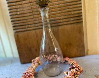 May include: A clear glass bottle with a dark brown glass stopper. The bottle is sitting on a light blue surface with a wreath of dried pink flowers around it.