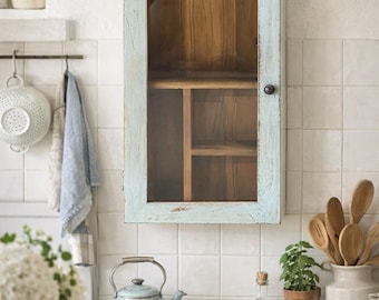 May include: A light blue, distressed-finish wall cabinet with a glass door and wooden interior. The cabinet is mounted on a white tiled wall. A light blue kettle, wooden spoons, and a small potted plant are on the countertop.
