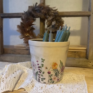 Könnte beinhalten: Ein weißer Keramiktopf mit floralem Design mit lila, rosa, gelben und grünen Blumen. Der Topf ist mit tealfarbenen Kerzen gefüllt.