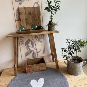 Könnte beinhalten: Ein rustikaler Holztisch mit dekorativen Glaskugeln in einer Holzschale, einem kleinen Bonsai-Baum und einem Vintage-Botanikausdruck. Ein grauer Filzteppich mit weißem Herzdesign und eine kleine Holzkiste ergänzen die Szene.
